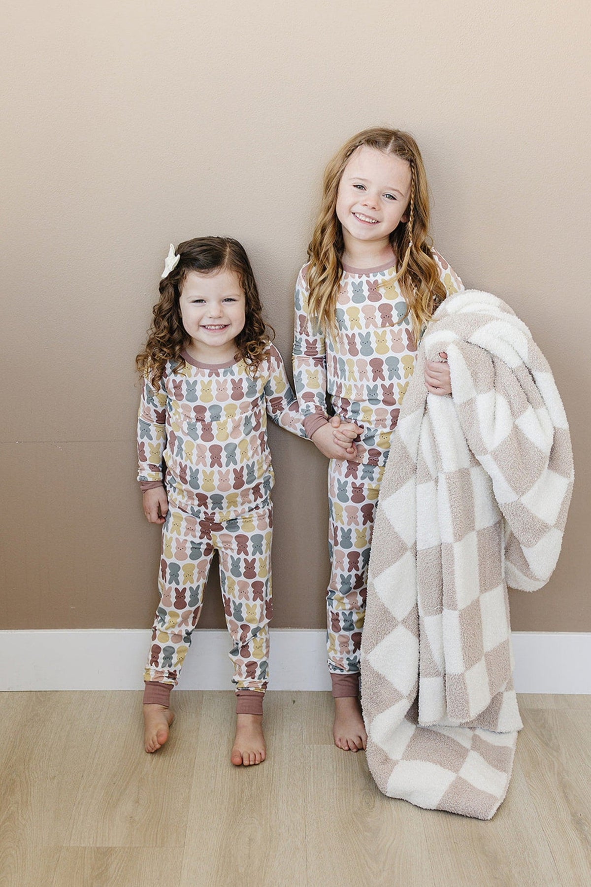 Two smiling children wearing matching bunny-patterned pajamas hold hands, with one holding a checkered blanket.