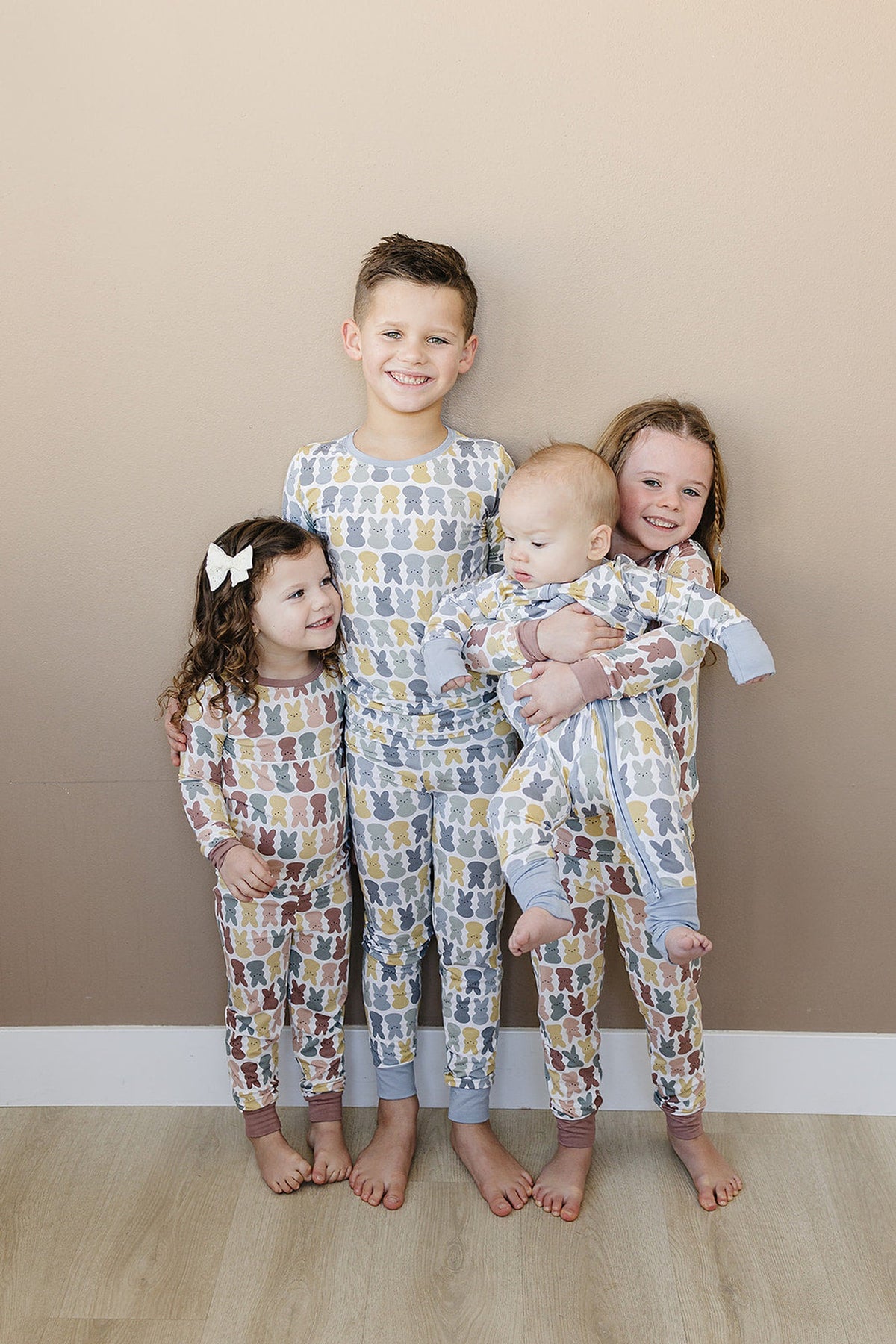 Four children in matching bunny-patterned pajamas stand against a beige wall, smiling and holding a baby.