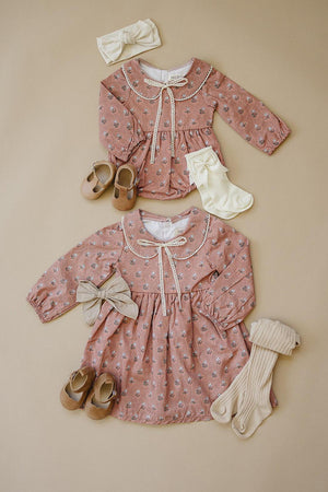 Two pink floral dresses with lace-edged collars, paired with bows, shoes, and socks on a beige background.