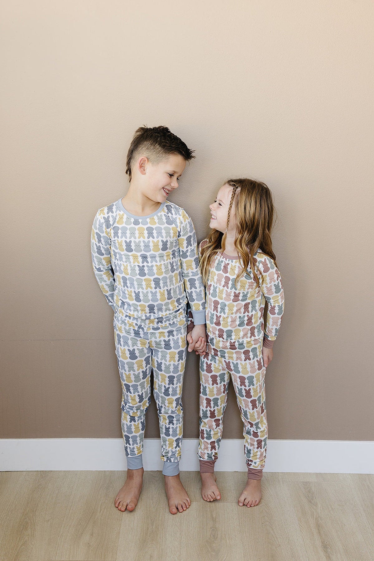 Two children smiling and holding hands, wearing matching bunny-patterned pajamas in pastel colors against a beige wall.