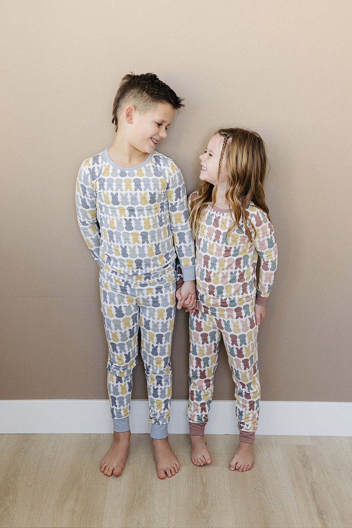Two children smile at each other wearing matching pajama sets with multicolored bunny patterns, standing barefoot indoors.