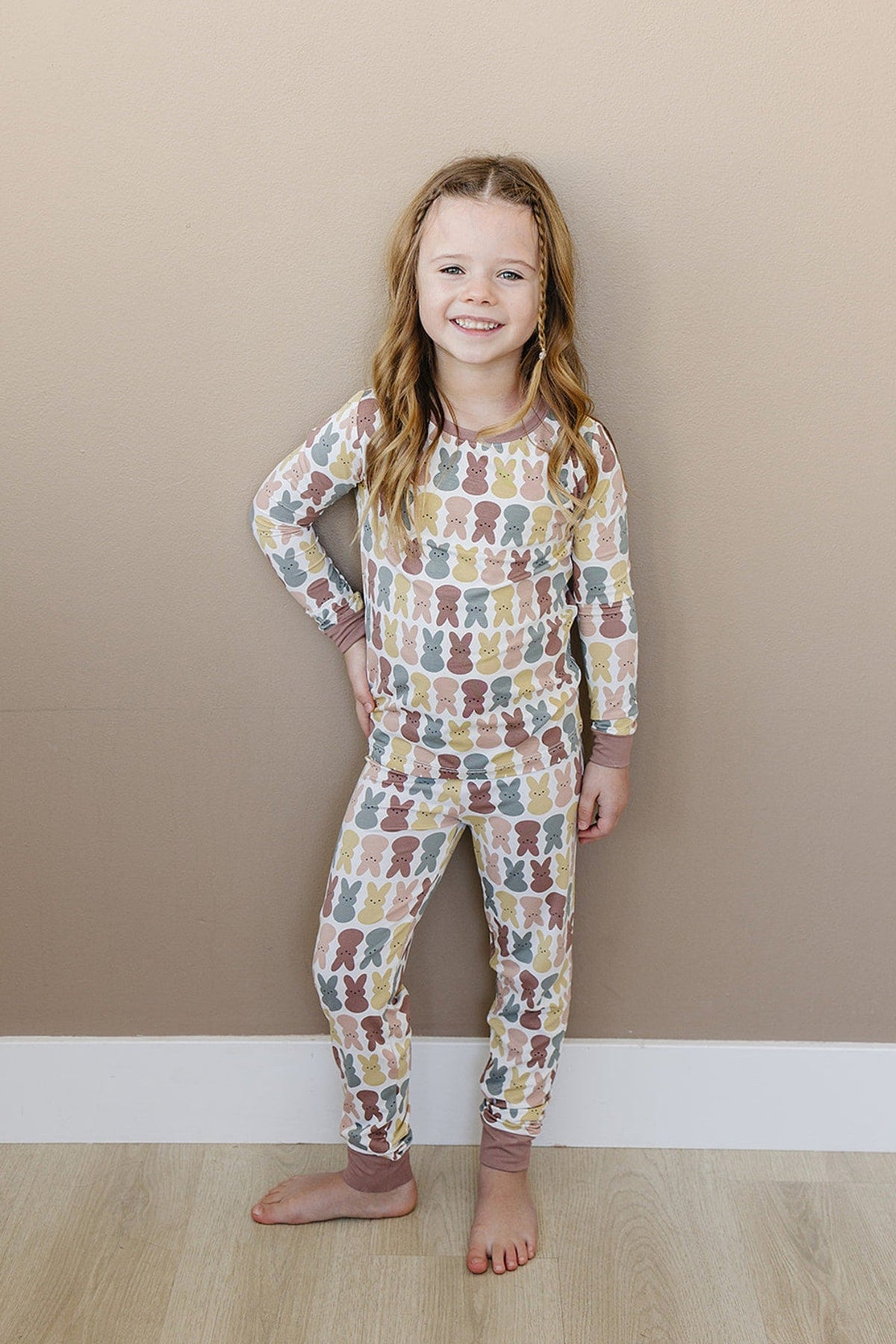 Young girl wearing a colorful bunny-patterned pajama set, standing against a beige wall, smiling with hands on hips.
