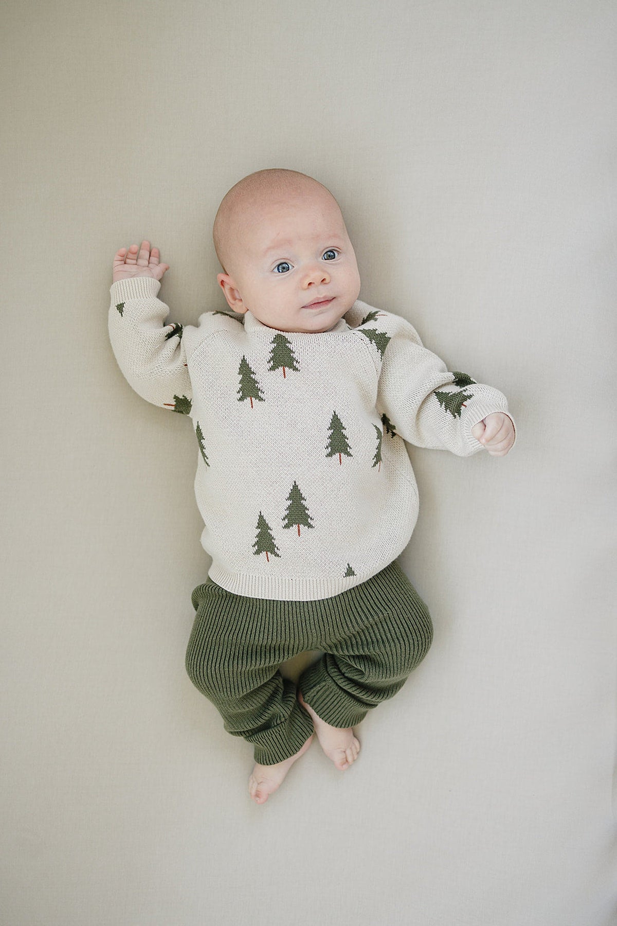A baby wearing olive knit leggings and a cream sweater with green tree patterns lies on a light surface.