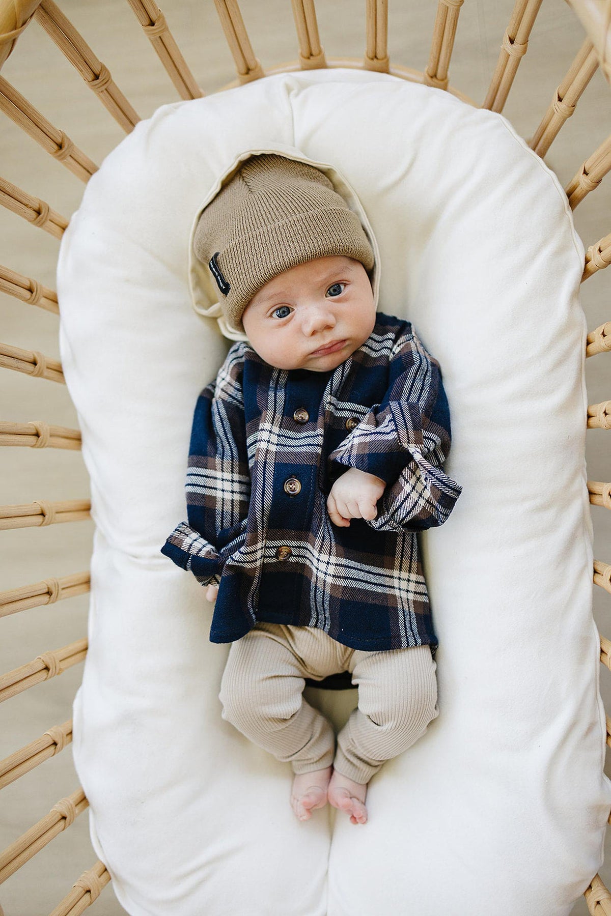 Baby in plaid button-up shirt and beige beanie lies on a white cushion in a wicker bassinet.