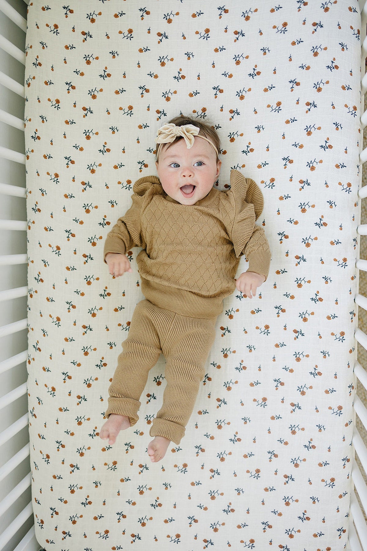 A baby in toffee knit leggings and sweater lies on a floral-patterned crib sheet, smiling.