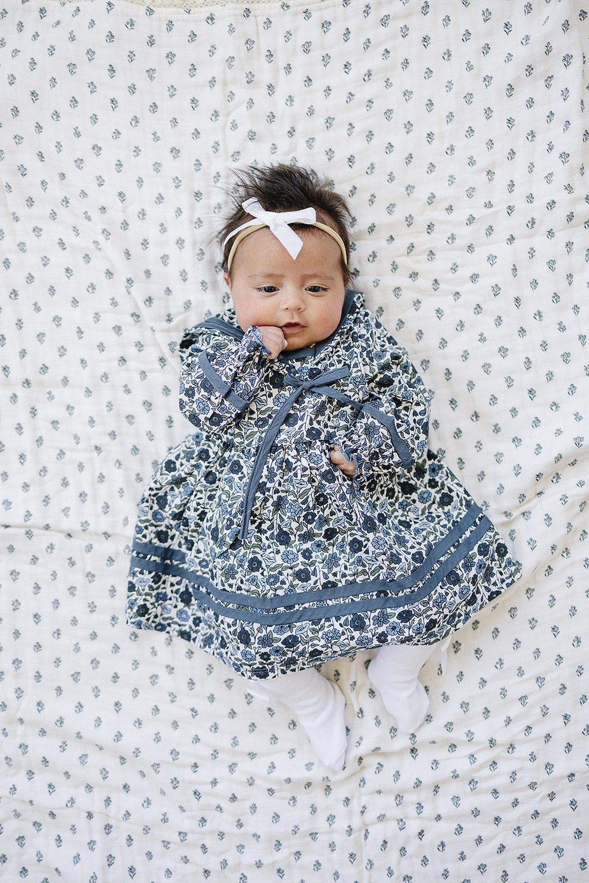A baby in a floral sailor dress with a bow headband lies on a patterned blanket.