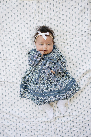 A baby in a floral sailor dress with a bow headband lies on a patterned blanket.