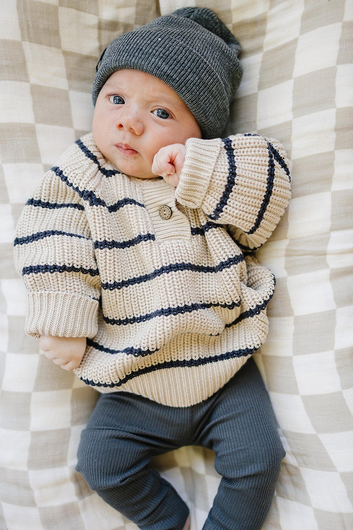 A baby wearing a beige and navy striped knit sweater and gray beanie lies on a plaid blanket.