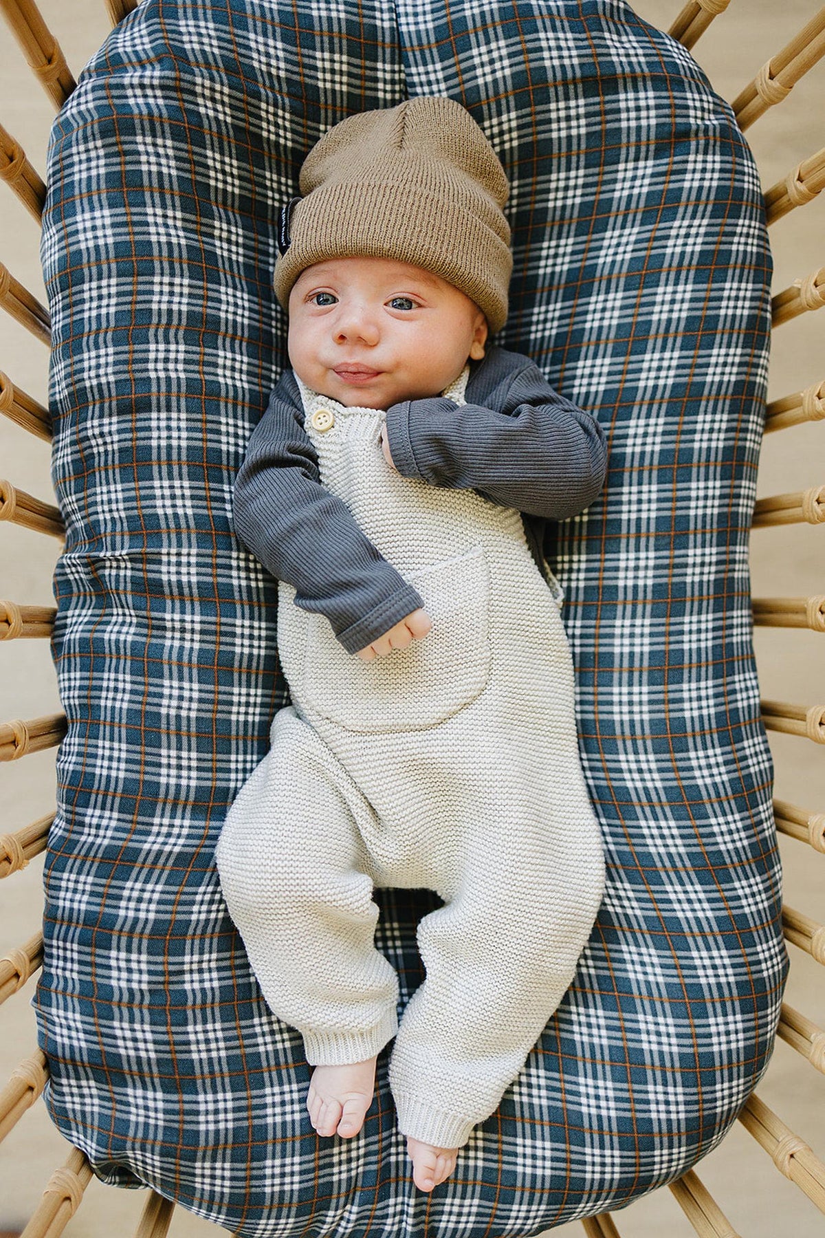 A baby wearing beige knit overalls and a brown beanie lies on a plaid-patterned cushion.