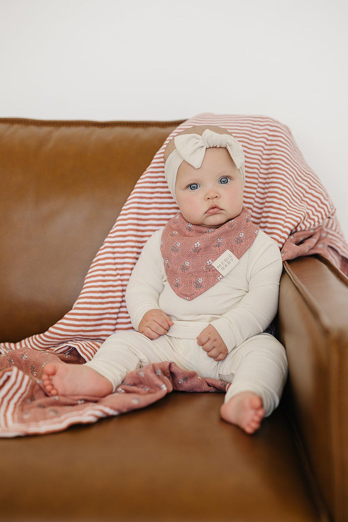 A baby wearing a cream outfit and headband sits on a brown couch with a striped blanket draped behind.