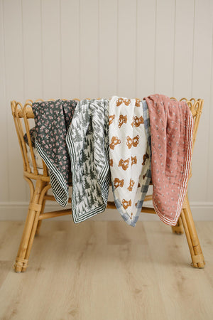Four muslin quilts with floral, forest, teddy bear, and pink stripe patterns draped over a bamboo crib.