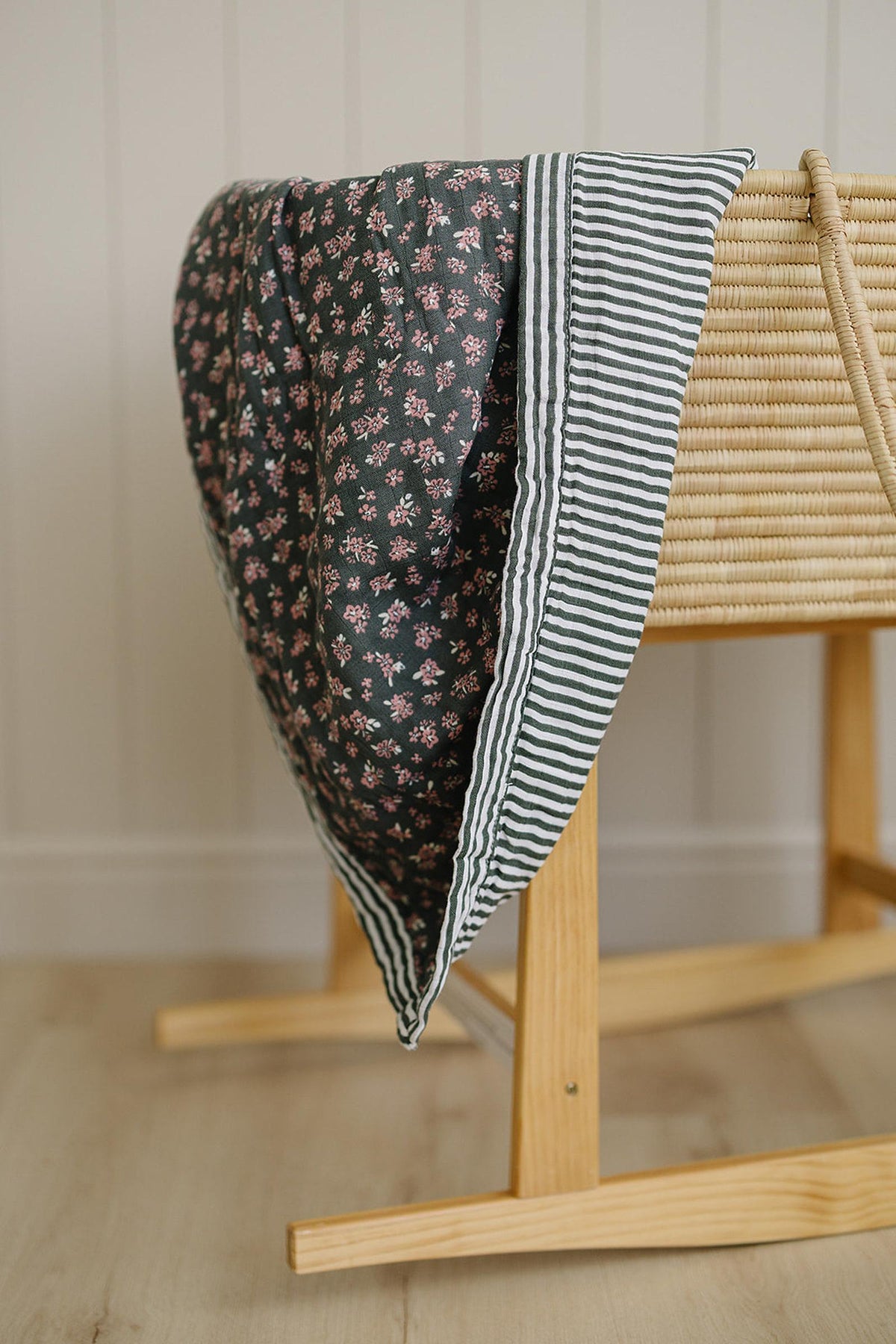 A floral and striped muslin quilt draped over a wooden crib, featuring a dark background and pink flowers.