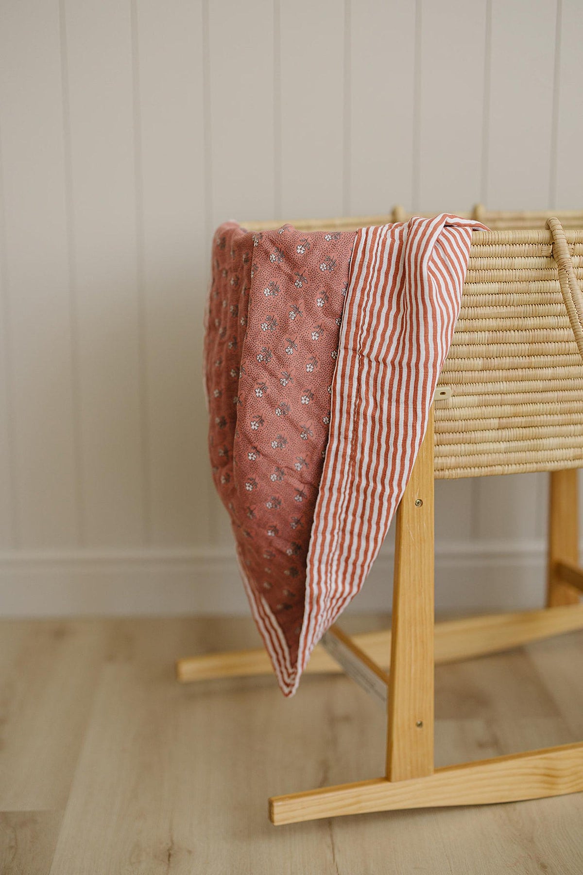 A pink floral and striped muslin quilt draped over a light wooden crib against a neutral wall.