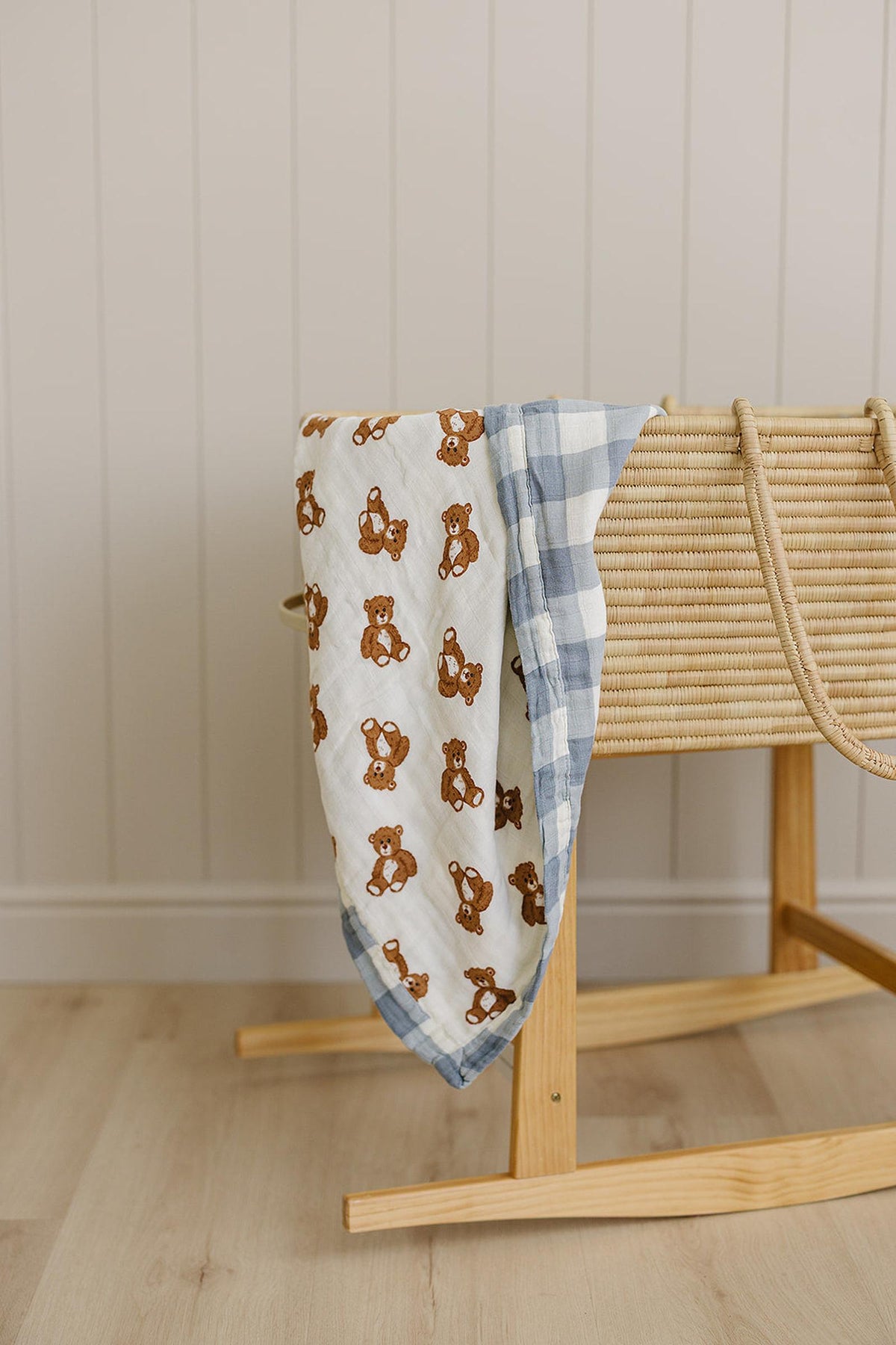 A muslin crib quilt featuring a teddy bear print and a dusty blue gingham pattern drapes over a wooden rocking bassinet, adding a cozy touch to a neutral nursery setting.