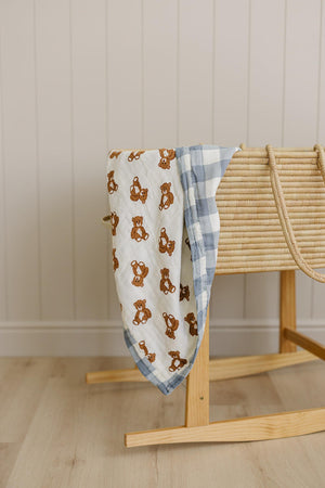 A muslin crib quilt featuring a teddy bear print and a dusty blue gingham pattern drapes over a wooden rocking bassinet, adding a cozy touch to a neutral nursery setting.