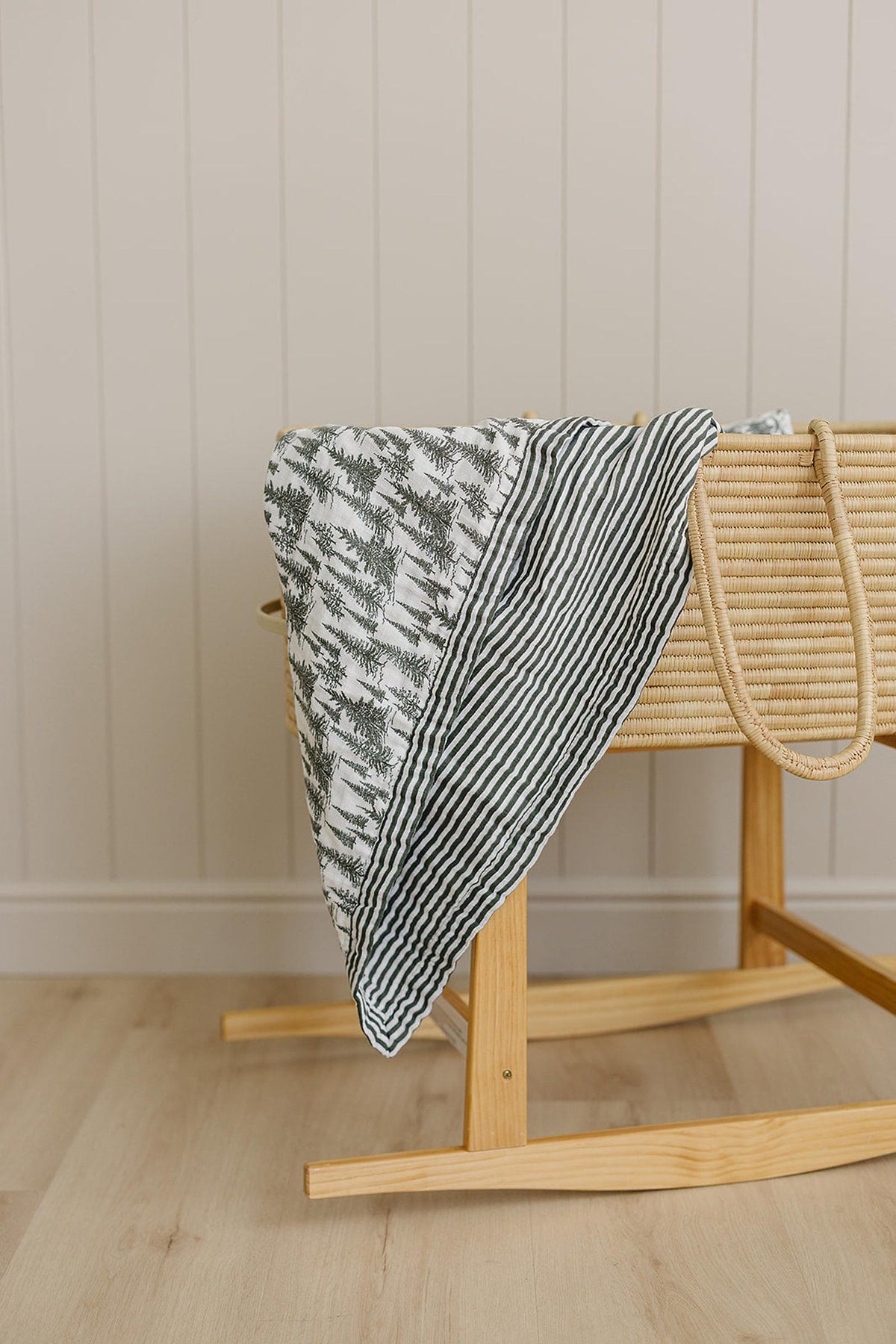 A muslin quilt with forest and green stripe patterns draped over a wicker bassinet in a nursery.