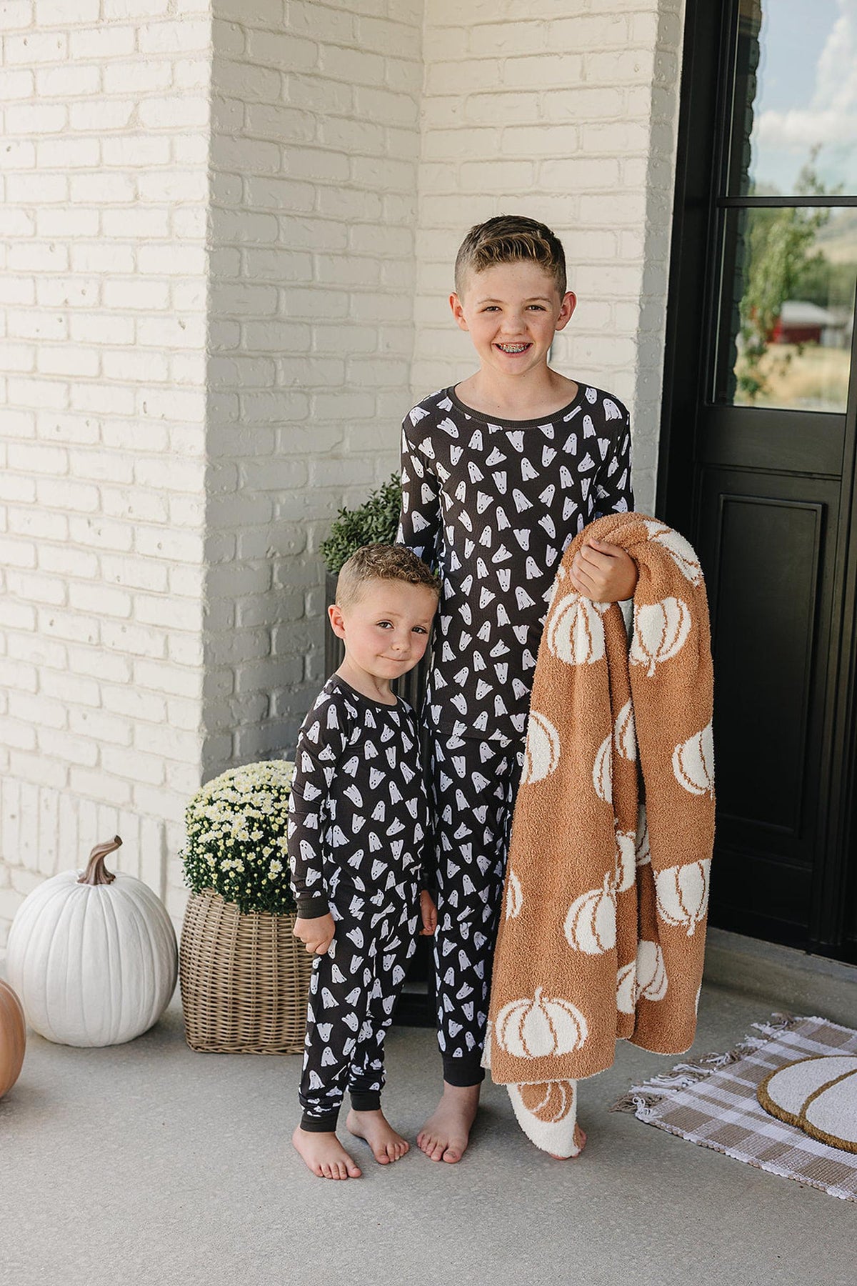 Two children wearing matching black pajamas with white ghost prints stand on a porch holding a pumpkin-patterned blanket.