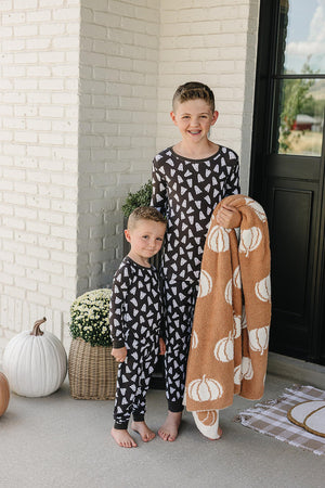 Two children wearing matching black pajamas with white ghost prints stand on a porch holding a pumpkin-patterned blanket.