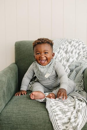 A smiling baby in a gray outfit sits on a green chair with a striped muslin quilt.
