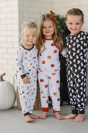Three children wearing festive Halloween-themed pajamas with pumpkin and ghost patterns, standing together on a porch.