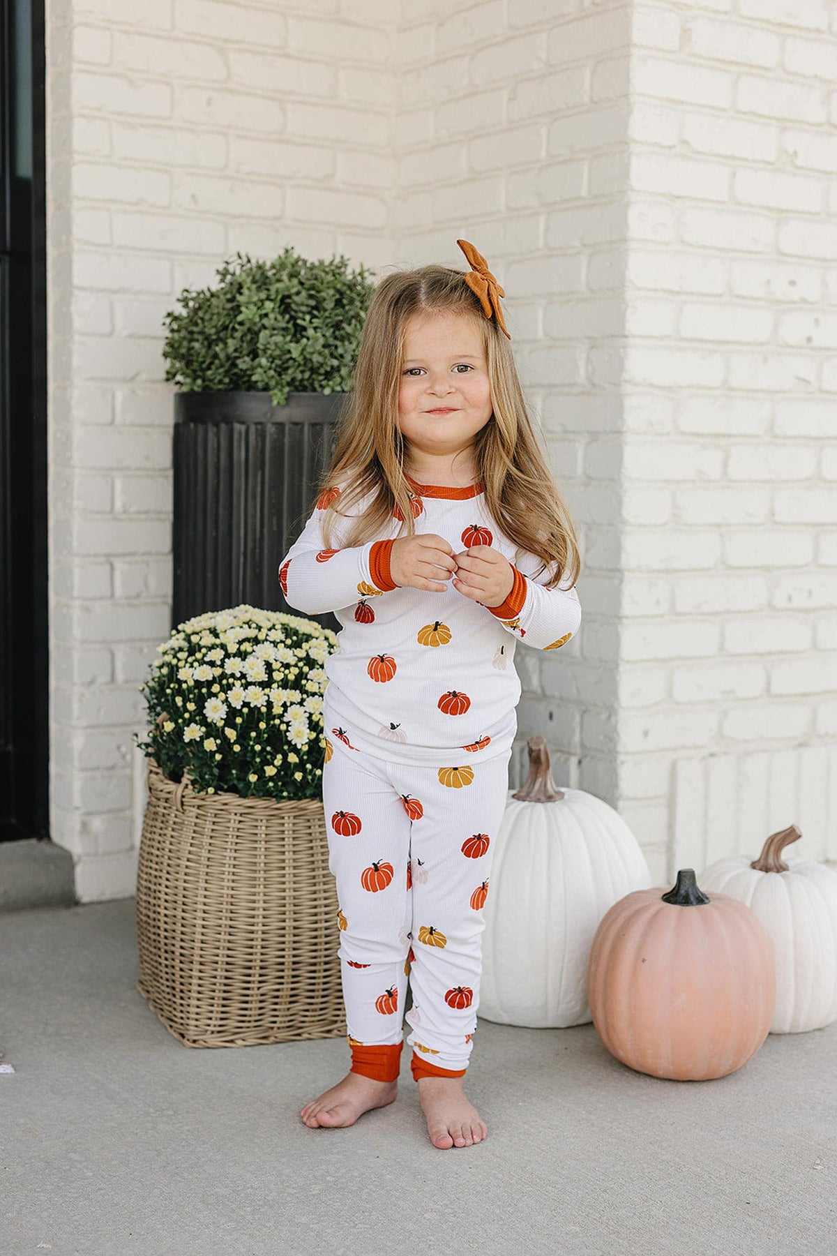 A child wearing a white pajama set with orange pumpkin prints stands barefoot near white pumpkins and a plant.