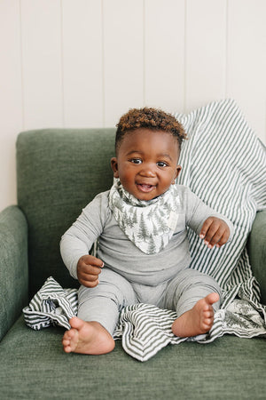 A happy baby wearing a forest-patterned muslin bib and gray outfit sits on a green chair with striped fabric.