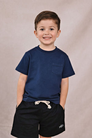 A smiling child wears a navy cotton T-shirt with a chest pocket, paired with black shorts, against a neutral backdrop.