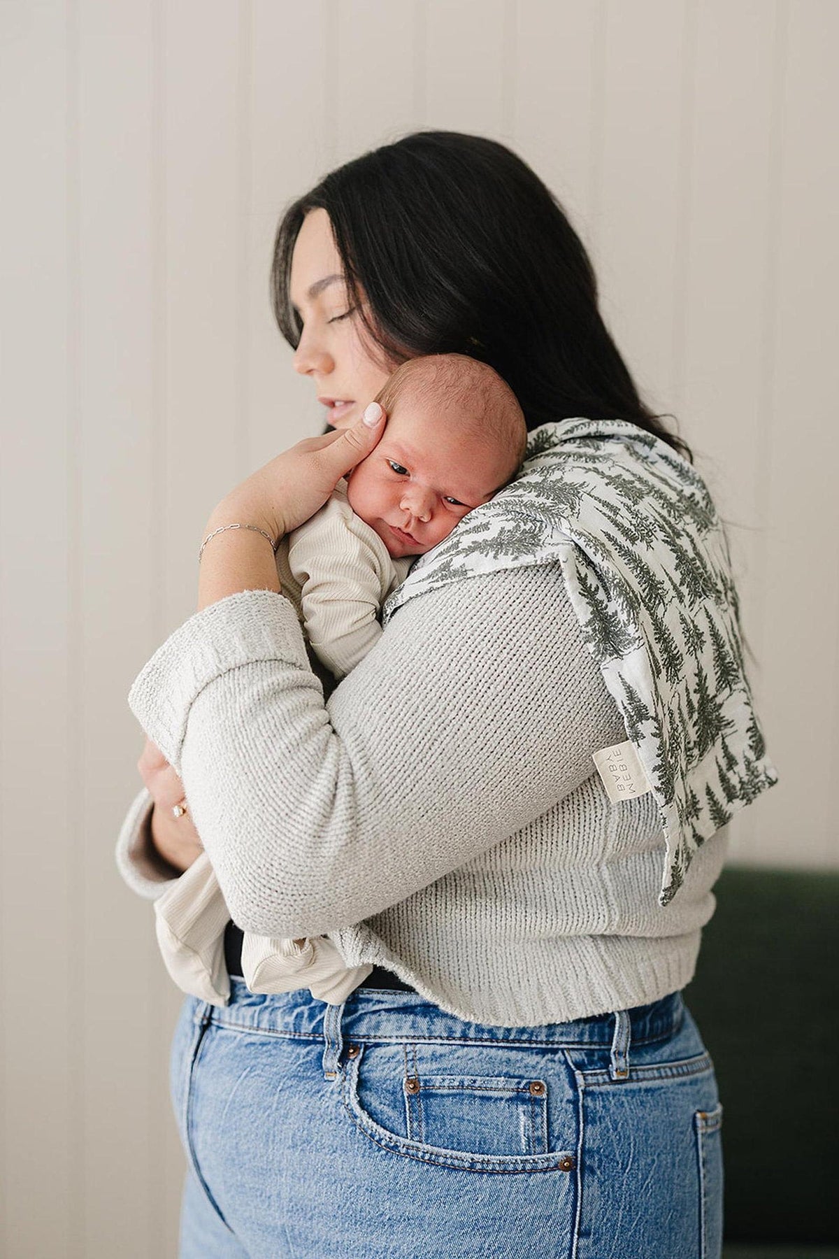 A woman holds a baby with a forest-patterned muslin burp cloth on her shoulder in a cozy setting.