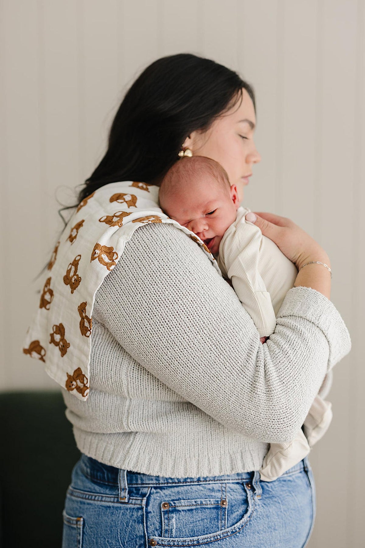 A woman holds a sleeping baby on her shoulder with a white muslin burp cloth featuring teddy bears.