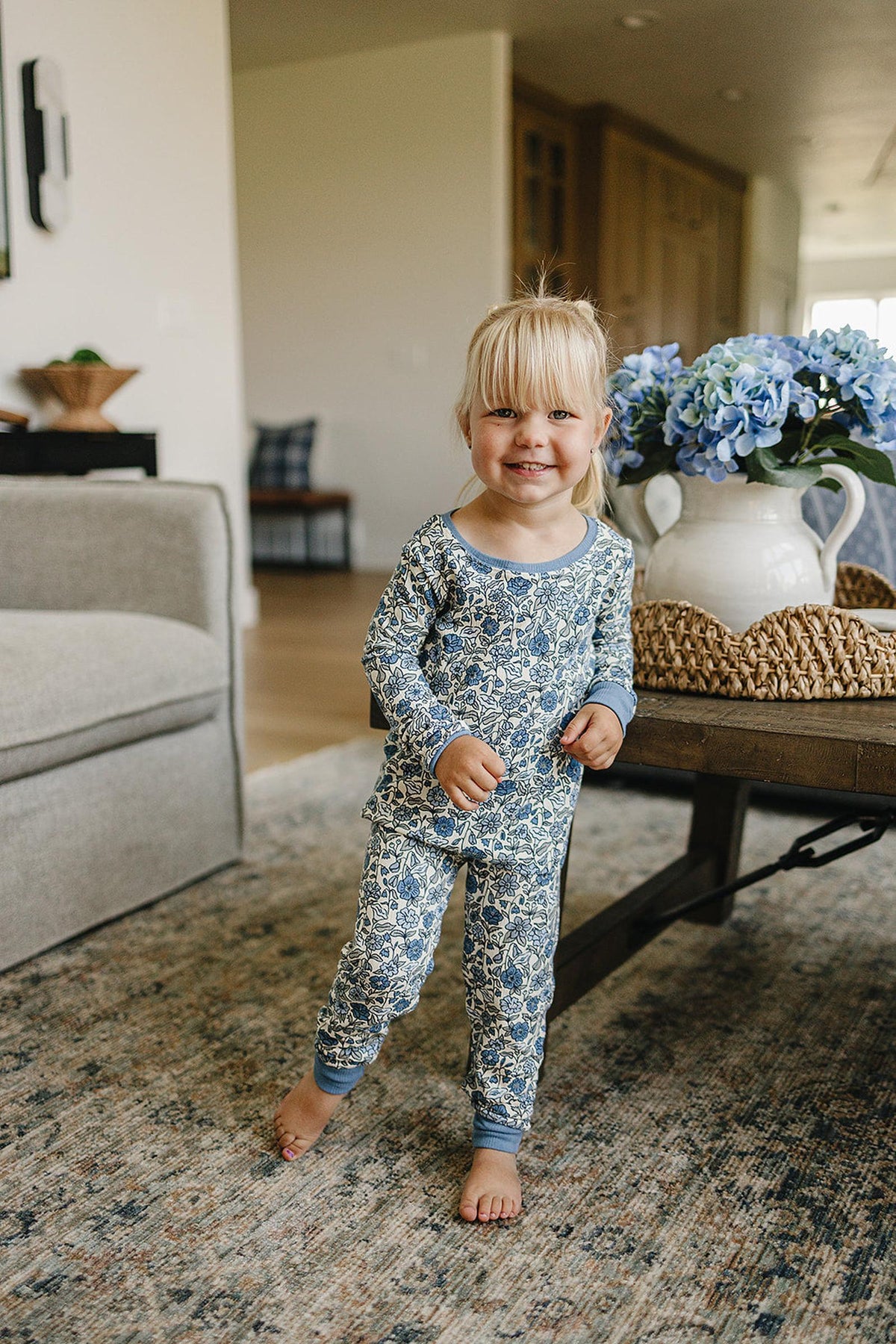 A child wearing a floral bamboo pajama set smiles in a cozy living room with a vase of blue flowers.