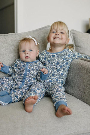 Two children in floral blue pajamas sit on a gray couch, smiling and looking content.
