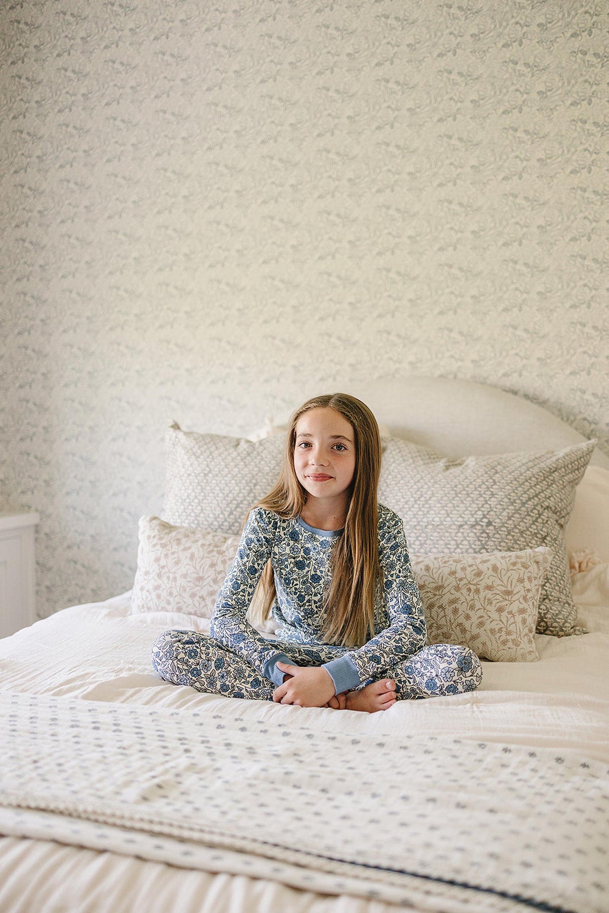 A child in a blue floral pajama set sits cross-legged on a bed with patterned pillows.