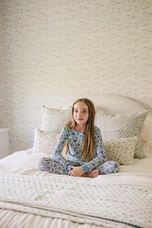 A child in a blue floral pajama set sits cross-legged on a bed with patterned pillows.