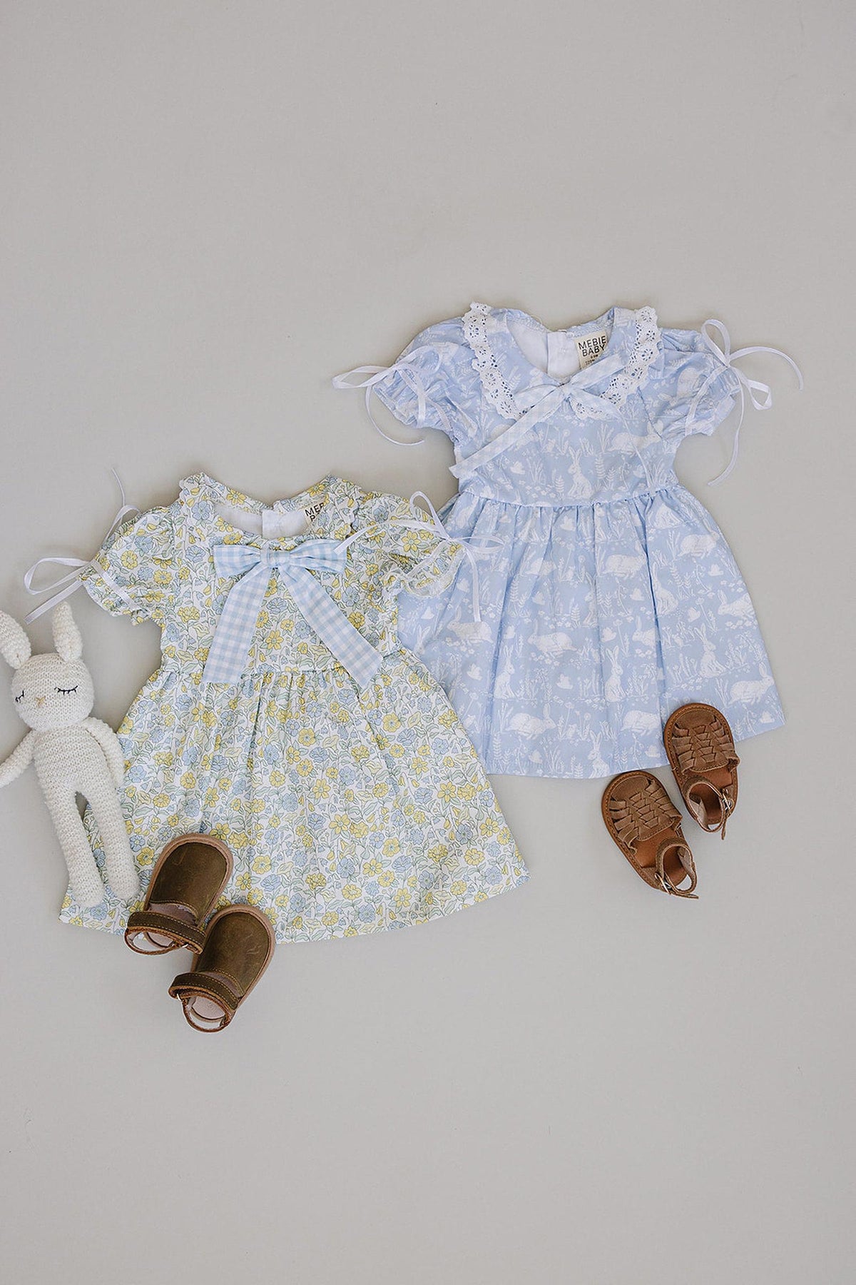Two adorable Mebie Baby floral dresses in yellow and light blue, featuring bows and lace details, paired with brown sandals and a plush bunny toy on a neutral background.