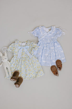 Two adorable Mebie Baby floral dresses in yellow and light blue, featuring bows and lace details, paired with brown sandals and a plush bunny toy on a neutral background.
