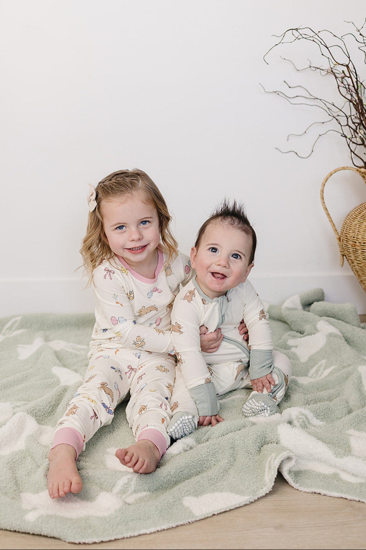 A girl and a baby sit on a soft, green blanket, wearing cozy pajama sets featuring playful bunny patterns.