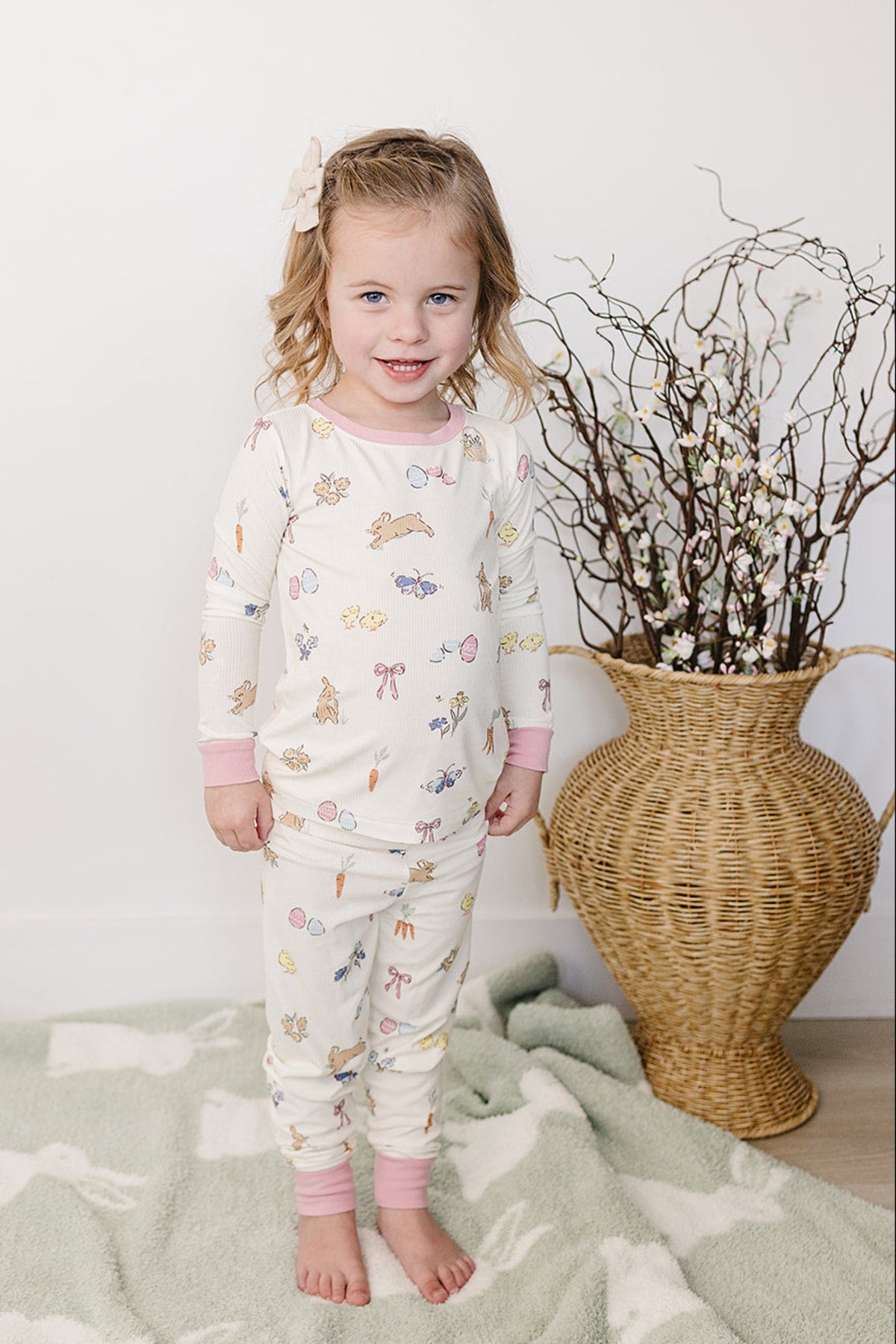 A young girl smiles while wearing a pastel-colored pajama set featuring playful bunny and floral patterns, standing on a soft.