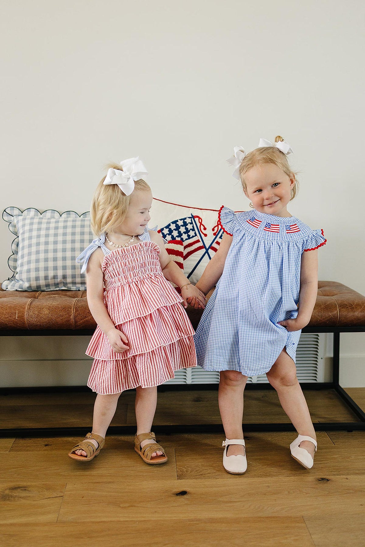 Two toddlers wearing patriotic dresses with white bows stand on a wooden floor, smiling in a cozy room.