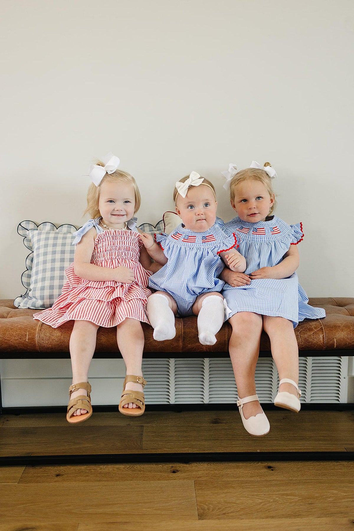 Three toddlers in red and blue striped dresses sit on a bench, each wearing white bows in their hair.