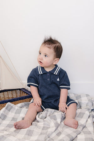 A baby boy sits on a checkered blanket, wearing a navy collared romper with white trim and an embroidered sailboat.