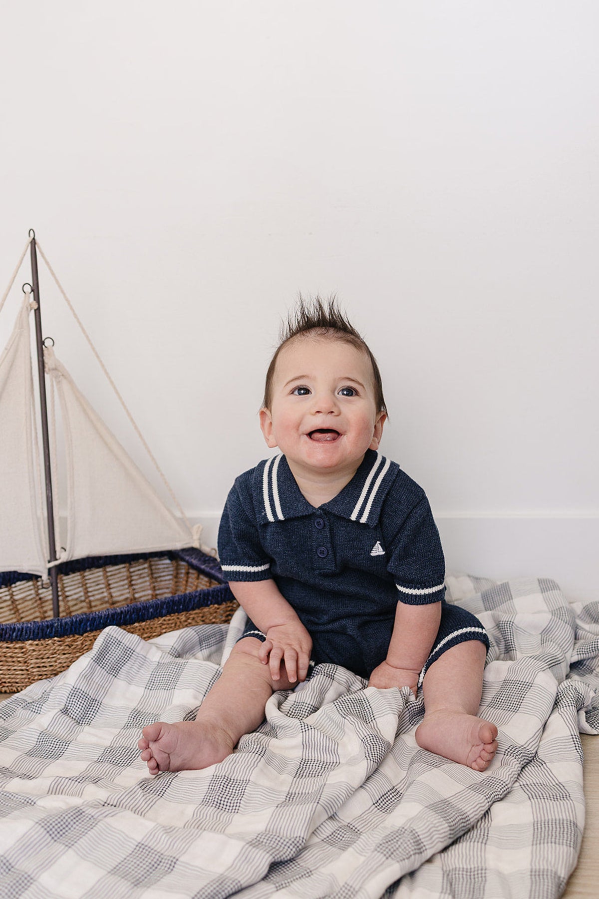 A smiling baby boy sits on a soft blanket, wearing a navy collared romper with white stripes, beside a small.
