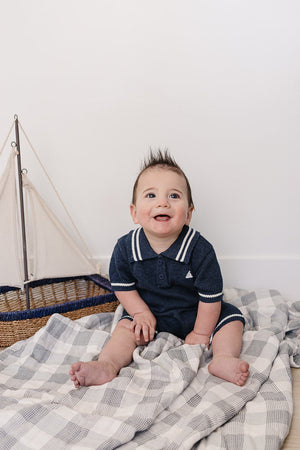 A smiling baby boy sits on a soft blanket, wearing a navy collared romper with white stripes, beside a small.