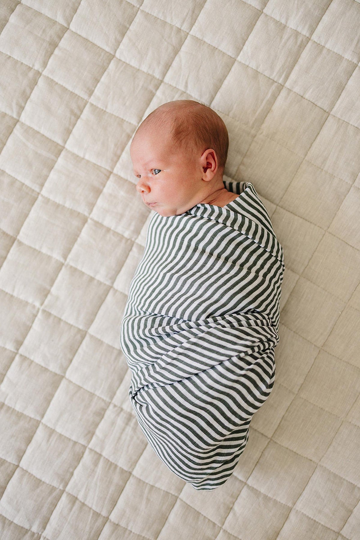 A baby wrapped in a green striped muslin swaddle blanket lies on a beige quilted surface.