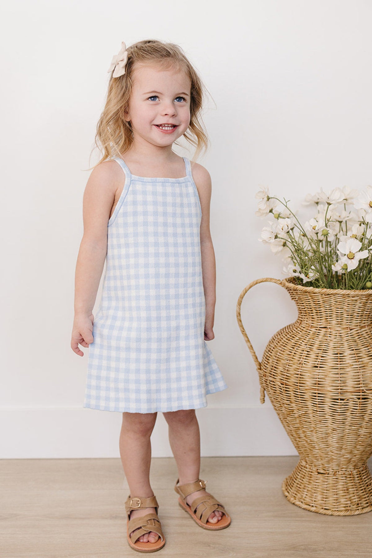 A young girl smiles while wearing a blue gingham dress with spaghetti straps, standing beside a woven vase of flowers.