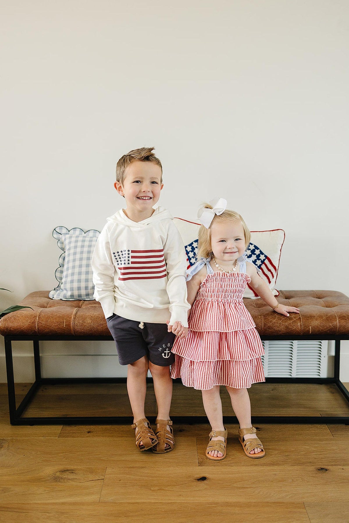 Two smiling children stand indoors; one wears a white hoodie with an American flag, the other a red striped dress.