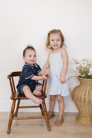 A smiling girl in a blue gingham dress stands beside a seated baby in a navy outfit on a wooden.