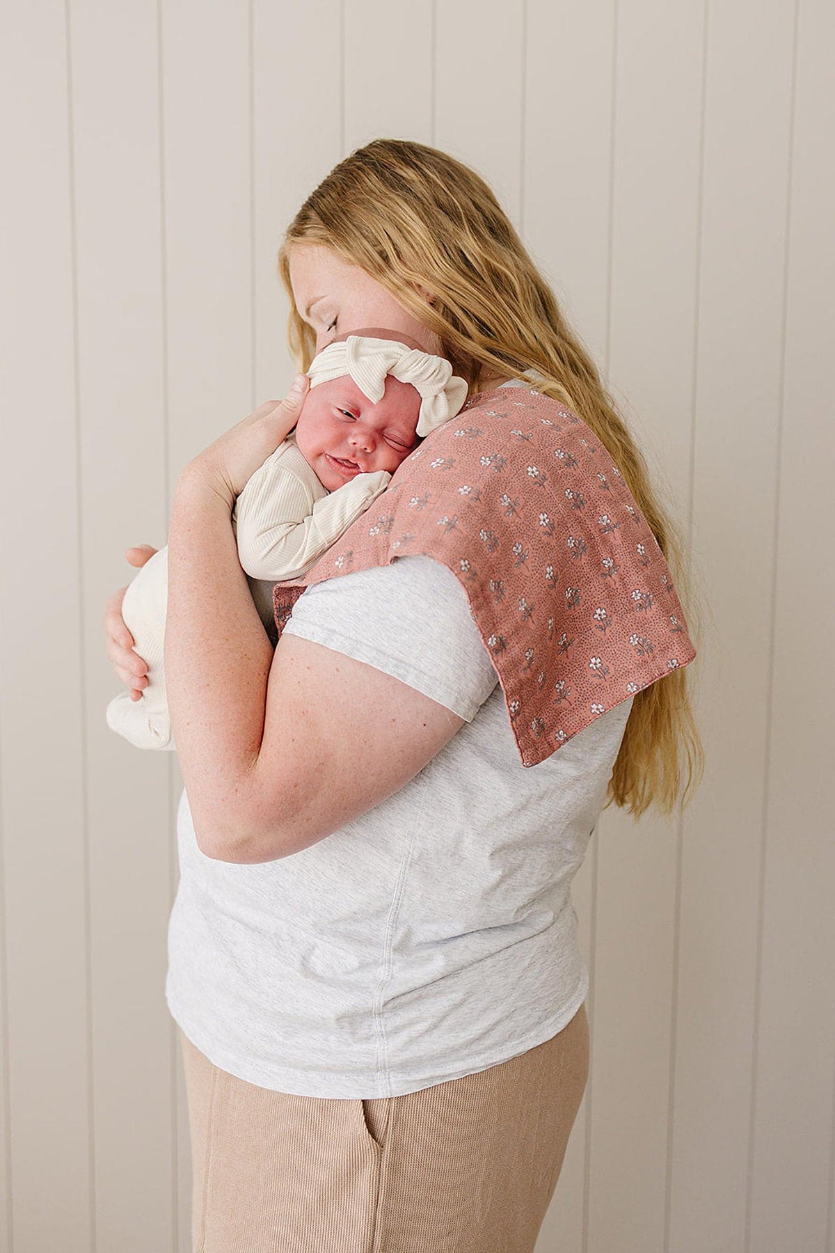 A woman gently holds a baby with a floral muslin burp cloth on her shoulder, set against a neutral wall.