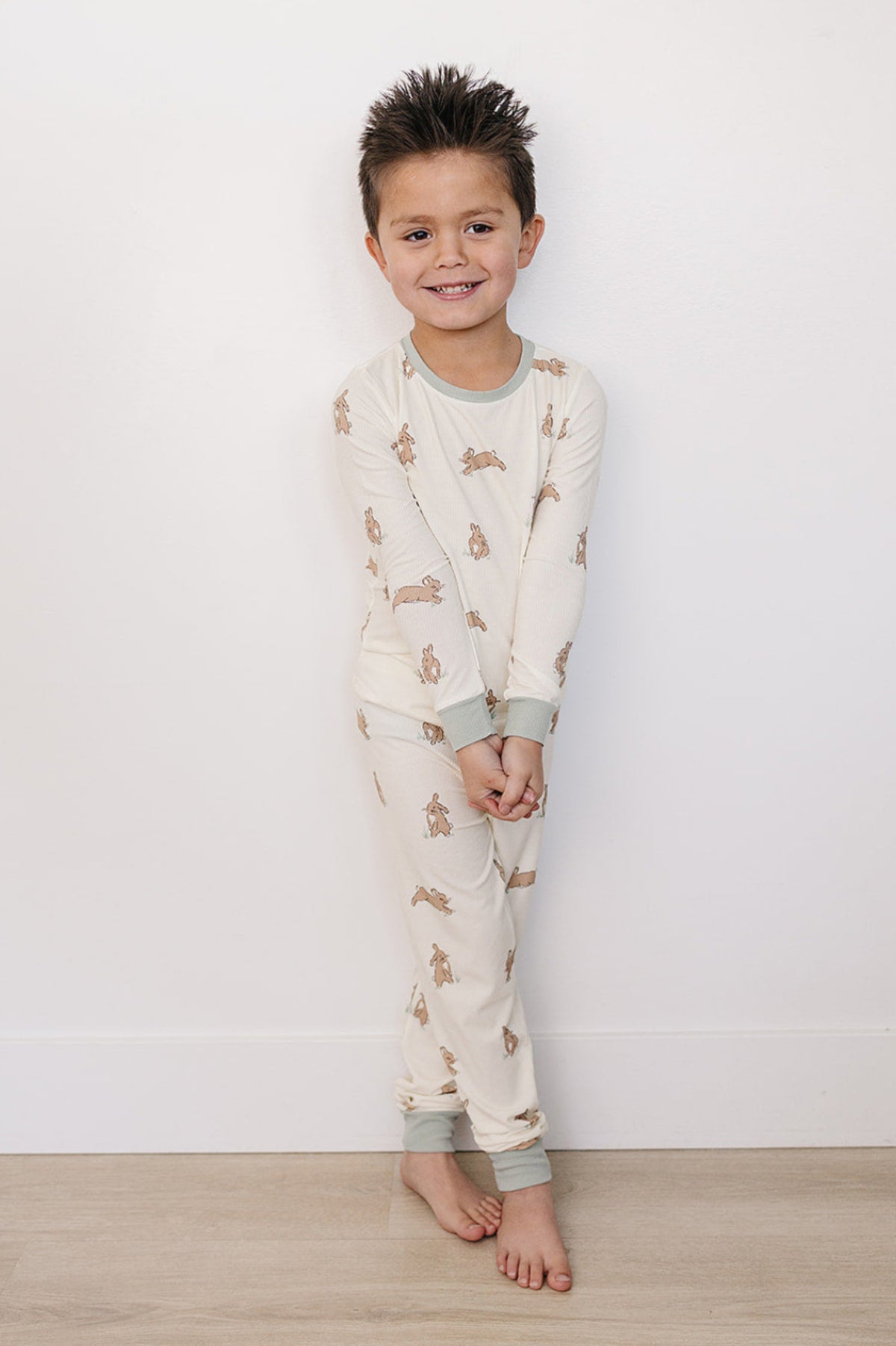 A smiling boy stands against a white wall, wearing a light cream pajama set featuring a playful bunny print, made from soft bamboo fabric, perfect for toddlers and kids.