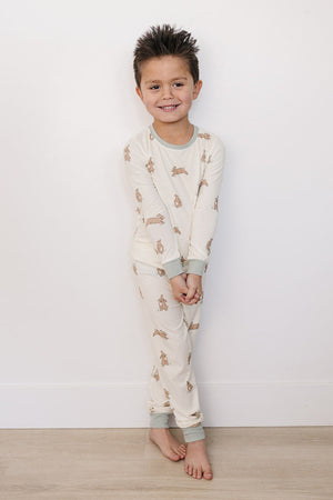 A smiling boy stands against a white wall, wearing a light cream pajama set featuring a playful bunny print, made from soft bamboo fabric, perfect for toddlers and kids.