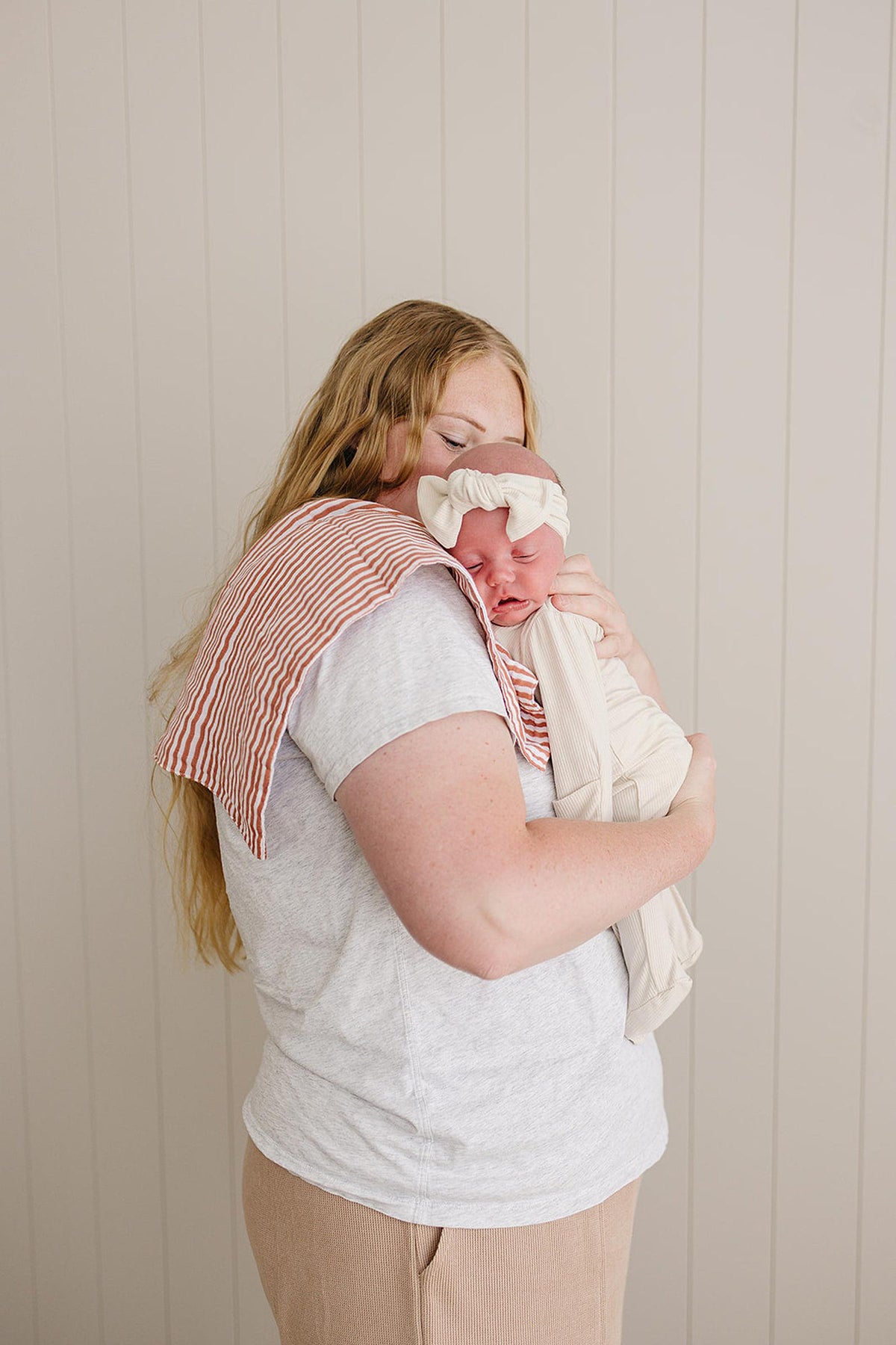 A woman holds a baby wrapped in cream fabric, with a pink striped burp cloth on her shoulder.